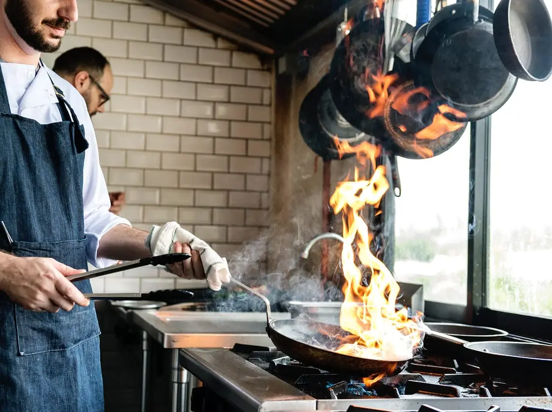 Chef flambés food in a pan beneath a commercial kitchen extraction hood